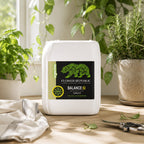 5 gallons of Flower Republic Balance:SI soil supplement sits on a wooden table surrounded by potted green plants and a cloth, with a window in the background.