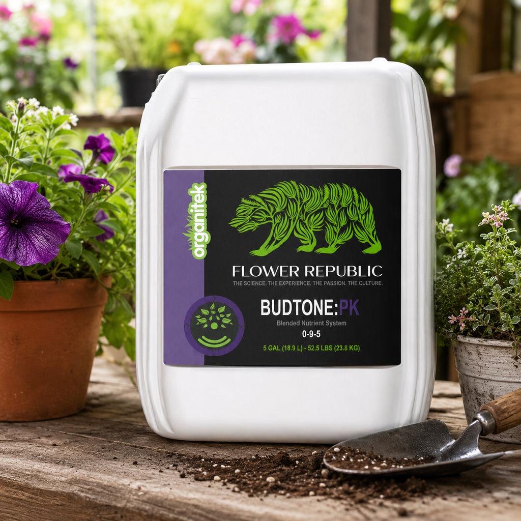 5 gallons of Flower Republic BudTone:PK blended nutrient system fertilizer, placed on a wooden table with soil and gardening tools, flanked by potted purple flowers in a garden setting.